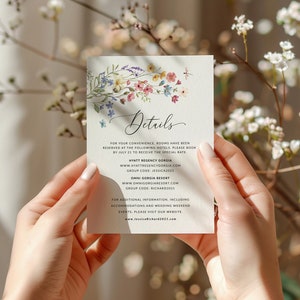 May include: A white card with a floral design and the word "Details" in cursive. The card provides hotel information for a wedding, including the hotel names, websites, and group codes. The text also includes a website address for additional information.