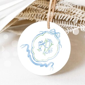 May include: A white round ceramic ornament with a blue and green watercolor monogram design. The monogram features the letters "A" and "V" intertwined with a blue ribbon and bow.