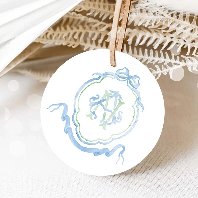 May include: A white round ceramic ornament with a blue and green watercolor monogram design. The monogram features the letters "A" and "V" intertwined with a blue ribbon and bow.