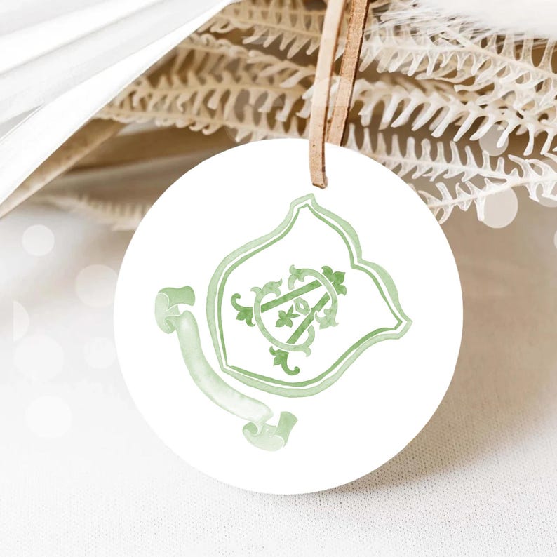 May include: A round, white ornament with a light green watercolor design. The design features a shield shape with intertwined initials and a banner. A brown leather string is attached for hanging. The background includes dried plants and a white surface.
