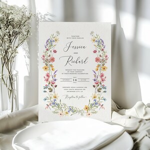 May include: A floral wedding invitation with a wreath of watercolor flowers in shades of pink, yellow, blue, and green. The invitation reads "Jessica and Richard" and includes the date, time, and location of the wedding.