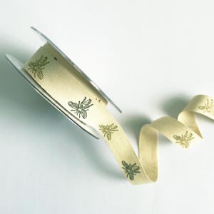 May include: A roll of beige cotton ribbon with a repeating pattern of gold bees. The ribbon is on a spool and a length of the ribbon is unfurled.