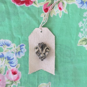 May include: A white fabric gift tag with a silver and clear rhinestone brooch in the shape of a crown. The tag is tied with a string and is on a green background with a floral pattern.