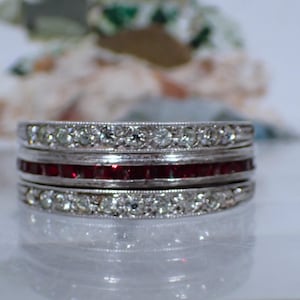 May include: A silver-toned ring featuring three stacked bands. The top and bottom bands are set with a row of clear, round-cut stones, while the center band contains a continuous row of square-cut, deep red stones set in a channel setting.