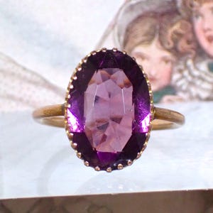 Op de afbeelding: Een gouden ring met een grote, ovaal geslepen paarse edelsteen in een geschulpte metalen zetting. De gefacetteerde steen vertoont diepe violette tinten en is bevestigd op een eenvoudige, dunne gouden ring met een gepolijste afwerking.