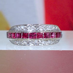 May include: A silver-toned ring featuring a central horizontal row of square-cut vibrant red rubies flanked by two parallel rows of small, clear, round-cut white stones. The band has a polished metallic finish and a structured, multi-row design.