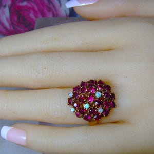 May include: A gold-toned ring featuring a cluster of small, round, deep red gemstones, interspersed with a few smaller, white opalescent stones. The ring is set on a light-toned surface, showcasing the intricate design and color contrast.