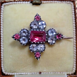 Pode incluir: Um broche vintage com um design em forma de cruz. O broche apresenta uma grande pedra preciosa rosa retangular no centro, rodeada por pedras preciosas claras e rosas. O broche é definido em metal prateado e é exibido em uma caixa forrada de veludo.