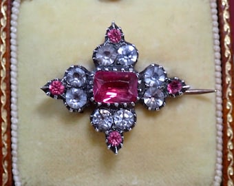 May include: A vintage brooch with a cross-shaped design. The brooch features a large, rectangular, pink gemstone at the center, surrounded by clear and pink gemstones. The brooch is set in a silver-toned metal and is displayed in a velvet-lined box.