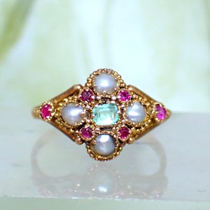 May include: A gold ring with an elaborate design. It has a central, square-cut, pale green gemstone, encircled by pearls and small, round, red gemstones. The ring's setting has a detailed, textured pattern.
