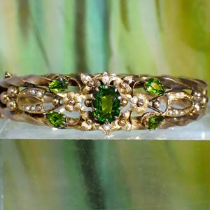 Op de afbeelding: Een goudkleurige scharnierende armband met een centrale ovaal geslepen groene edelsteen, omringd door kleine witte parels en vier peervormige groene stenen. De metalen band heeft een gedraaid ontwerp met florale accenten en textuur.