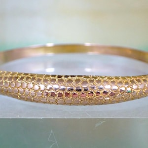 Puede incluir: Un brazalete rígido articulado de tono dorado con un patrón texturizado similar a un panal en su superficie exterior. La pulsera tiene un interior pulido e incluye una pequeña cadena de seguridad unida al mecanismo de cierre lateral.