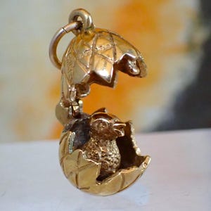 May include: A gold-coloured charm depicting an egg with an open top, revealing a small chick inside. The egg has a textured surface and is suspended from a ring. This charm is likely a pendant or jewellery item.