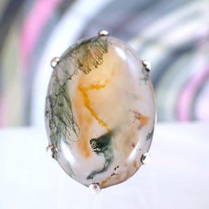 May include: Oval-shaped gemstone ring with a silver setting. The stone features a mix of colors, including white, green, and yellow, with a unique, natural pattern. The ring is a piece of jewelry.