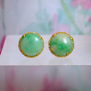 Op de afbeelding: Een paar ronde oorstekers met lichtgroene jade stenen in goudkleurige, getextureerde bezels. De oorbellen worden gepresenteerd op een helder, reflecterend oppervlak.