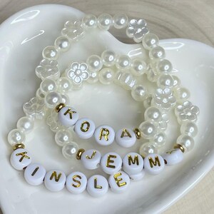 May include: Two elastic bracelets made of white faux pearl beads and white flower-shaped beads. One bracelet features white round beads with gold lettering spelling KORA, and the other features white round beads with gold lettering spelling KINSLEY JEMM.