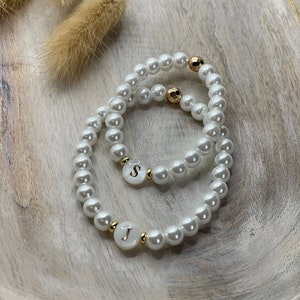 Puede incluir: Dos pulseras de perlas blancas con detalles dorados y abalorios con letras. Los abalorios deletrean las letras "J" y "S".