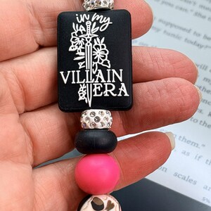 Puede incluir: Un cordón de cuentas con un dije rectangular negro que tiene el texto blanco in my VILLAIN ERA y un gráfico de espada. El accesorio incluye cuentas redondas de color rosa intenso, cuentas negras mate y cuentas plateadas con brillantes.