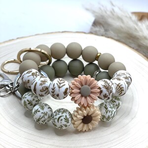 May include: A beaded keychain wristlet with olive green, tan, and white beads. The white beads have a botanical design. Peach and tan flower-shaped charms add detail. Includes gold and silver hardware. The wristlet is designed to be worn on the wrist.