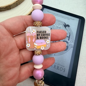Puede incluir: Un llavero de cuentas con un dije cuadrado que dice "Fueled by Coffee & Books". El llavero presenta cuentas rosas, lavanda y crema, detalles dorados y un dije dorado. El dije tiene un gráfico de taza de café y libros.