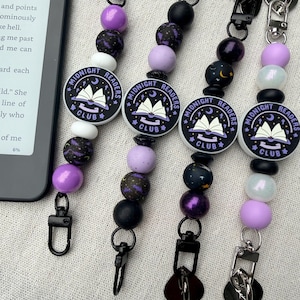 May include: Four beaded lanyards featuring black lobster clasps and round silicone beads in shades of purple, black, and white. Each lanyard includes a circular charm printed with the text Midnight Readers Club and an illustration of an open book.