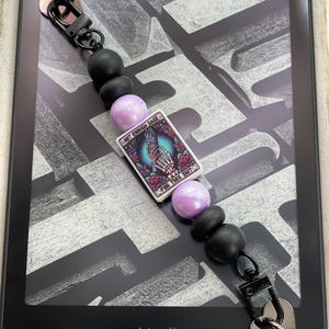 May include: A beaded accessory featuring matte black and pearlescent lavender beads, with a central rectangular charm depicting a skeleton hand holding a sage bundle, surrounded by roses and the text NOT ENOUGH SAGE FOR THIS SHIT, with black metal clasps.