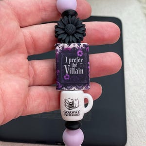 May include: A beaded lanyard with a black cord, featuring a black flower, a miniature book that says "I prefer the Villain", and a white mug that says "Go Away I'm Reading". The lanyard has white, lavender, and black beads.