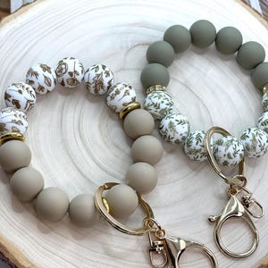 May include: Two beaded keychains. One keychain features taupe and white beads with gold floral designs, and the other has olive green and white beads with gold floral designs. Both have gold-colored hardware, including a clasp and a ring.
