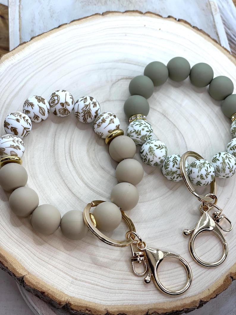 May include: Two beaded keychains. One keychain features taupe and white beads with gold floral designs, and the other has olive green and white beads with gold floral designs. Both have gold-colored hardware, including a clasp and a ring.