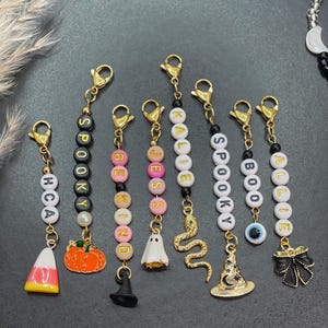 Op de afbeelding: Een verzameling Halloween-thema kralen sleutelhangers met goudkleurige sluitingen. De sleutelhangers hebben verschillende bedels, waaronder een pompoen, een spook, een slang en een heksenhoed. Sommige sleutelhangers spellen woorden zoals "SPOOKY".