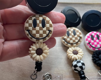 May include: A set of four retractable badge reels with different smiley face designs. The reels feature a black retractable cord, a clear plastic badge holder, and a decorative charm. The charms include a brown and white checkered smiley face, a white sunflower, a black and white checkered circle, and a pink and white checkered smiley face.