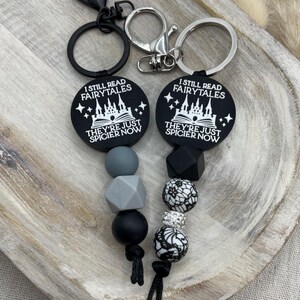 Peut inclure: Porte-cl&eacute;s &agrave; perles en silicone noir et gris avec deux porte-cl&eacute;s noirs. Les porte-cl&eacute;s pr&eacute;sentent un motif noir et blanc d'un ch&acirc;teau et le texte "I still read fairytales they're just spicier now".