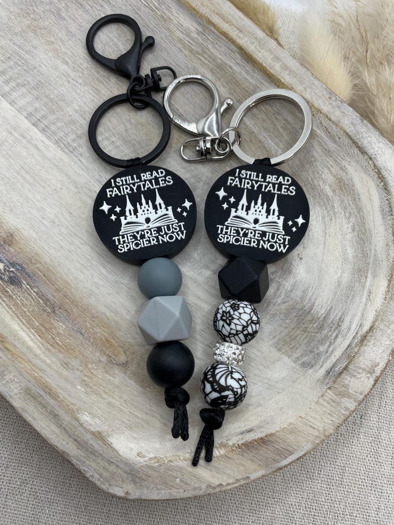 Peut inclure: Porte-cl&eacute;s &agrave; perles en silicone noir et gris avec deux porte-cl&eacute;s noirs. Les porte-cl&eacute;s pr&eacute;sentent un motif noir et blanc d'un ch&acirc;teau et le texte "I still read fairytales they're just spicier now".