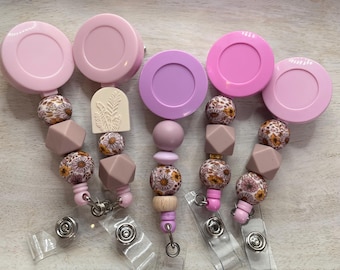 May include: Four pink and purple retractable badge reels with floral and geometric patterned beads. Each reel has a clear plastic badge holder.