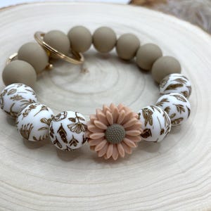May include: A beige silicone bead bracelet with a gold-colored keyring. The bracelet features a mix of solid beige and white beads with a brown floral pattern. A peach-colored flower is centered on the bracelet.