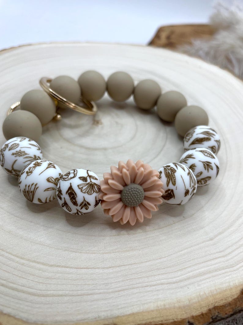 May include: A beige silicone bead bracelet with a gold-colored keyring. The bracelet features a mix of solid beige and white beads with a brown floral pattern. A peach-colored flower is centered on the bracelet.