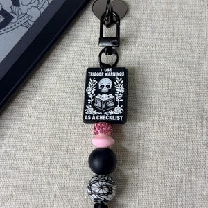 May include: A black keychain featuring a rectangular charm with a white skeleton reading a book, surrounded by floral motifs and the text I USE TRIGGER WARNINGS AS A CHECKLIST. Below are four beads: one pink rhinestone, one solid pink, one matte black, and one white with black lace patterns.