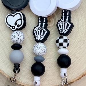 Może przedstawiać: Trzy chowane uchwyty na identyfikatory z różnymi wzorami. Jeden przedstawia serce ze stetoskopem, drugi znak pokoju ze szkieletem, a trzeci czarno-białą szachownicę. Koraliki są w kolorach czarnym, białym i szarym.