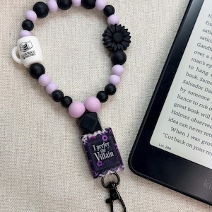 May include: A beaded lanyard in black, lilac, and white, with a black flower and a small mug that says "Go Away I'm Reading." It also features a charm that says "I Prefer the Villain" and a black clasp. The lanyard is next to an e-reader.