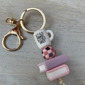 May include: A gold-coloured keyring with a lobster clasp and a large ring. The keyring features a white mug with "Read more books" text, a checkered bead, and two small book charms in shades of pink.