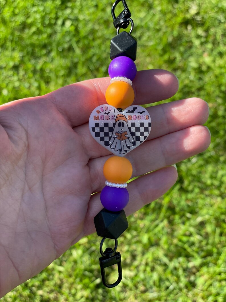 Pu&ograve; includere: Un portachiavi con perline a tema Halloween con chiusura nera. Il portachiavi presenta perline geometriche nere, perline rotonde viola e arancioni e un ciondolo a forma di cuore con un fantasma che legge un libro e la scritta "READ MORE BOOKS".