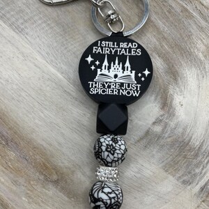 Peut inclure: Porte-cl&eacute;s noir et blanc avec un charme qui dit "I still read fairytales they're just spicier now". Le charme repr&eacute;sente un ch&acirc;teau et un livre ouvert. Le porte-cl&eacute;s comprend &eacute;galement des perles noires et blanches et un anneau de cl&eacute; argent&eacute;.