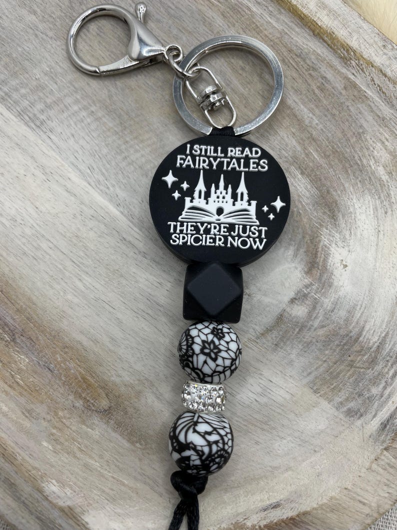 Peut inclure: Porte-cl&eacute;s noir et blanc avec un charme qui dit "I still read fairytales they're just spicier now". Le charme repr&eacute;sente un ch&acirc;teau et un livre ouvert. Le porte-cl&eacute;s comprend &eacute;galement des perles noires et blanches et un anneau de cl&eacute; argent&eacute;.