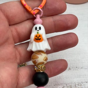 Może przedstawiać: Brelok o tematyce Halloween z białym duchem trzymającym dynię i pomarańczowym zapięciem w kształcie serca. Brelok zawiera ozdobne koraliki w kolorze kremowym, złotym, czarnym i różowym.
