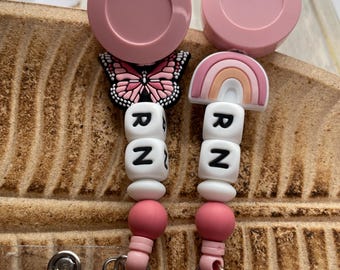 May include: Two pink retractable badge reels with clear badge holders. Each reel has a charm, one is a pink butterfly and the other is a rainbow. Both reels have white beads with the letters "RNZ" on them.