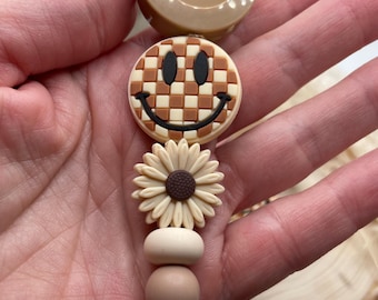 May include: A brown and white checkered smiley face charm with a brown daisy charm and brown wooden beads on a retractable badge reel.