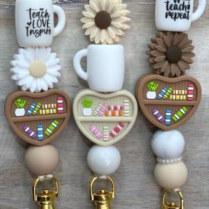 May include: Three beaded lanyards with gold-coloured clasps. Each lanyard features a white mug with text, a flower, a heart-shaped bookshelf, and various beads. The mugs say "Teach Love Inspire" and "coffee teach repeat".