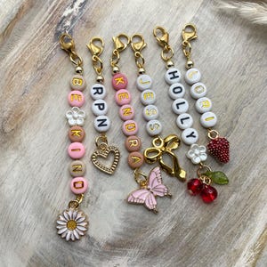 May include: Five colourful beaded keychains with gold clasps and charms. The keychains spell out the names "Lori", "Holly", "Jess", "Kendra", and "Breckin". Each keychain has a different charm, including a strawberry, cherries, a butterfly, a bow, and a heart.