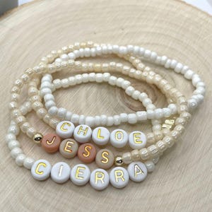 May include: A set of three beaded bracelets with the names "Chloe", "Jess", and "Cierra" spelled out in gold letters on white beads. The bracelets are made with white and cream-colored beads.