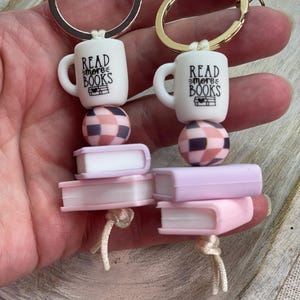 May include: Two keychains with gold-colored rings. Each keychain features a white mug with "Read More Books" text, a checkered bead, and a stack of pink and white books. The keychains are connected with a white cord.